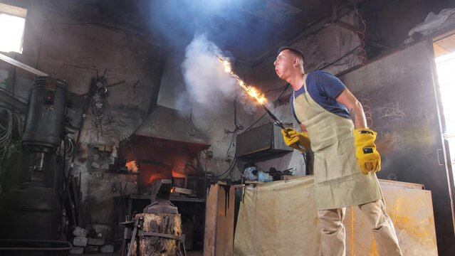 A blacksmith in a protective apron in the forge holds a forged sword burning in his hands, after quenching in oil.
