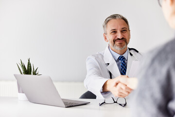 A senior doctor sitting in front of the laptop, and shaking hands with a patient.