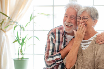 Happy senior couple embracing and laughing sitting at home. Modern retirees laugh at something...