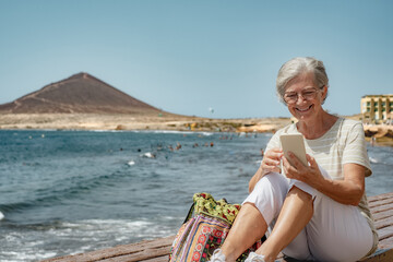 Fototapeta premium Video call concept. Smiling senior woman using smartphone for online communication via video meeting sitting at the beach, happy grey-haired woman looking and talking into phone webcam