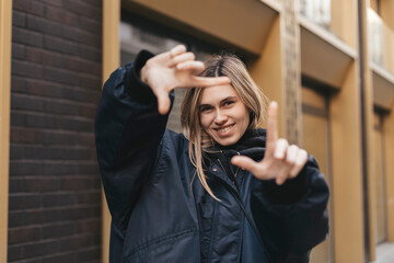 Attractive woman with a lovely smile making a frame gesture with her fingers framing her face. Woman posing on selfie photo looking at camera walking outdoors in urban city.