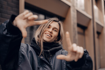 Fototapeta premium Attractive woman with a lovely smile making a frame gesture with her fingers framing her face. Woman posing on selfie photo looking at camera walking outdoors in urban city.