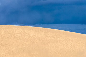 View of desert sand dunes against a moody cloudy blue sky. Maspalomas Dunes in Playa del Ingles,...