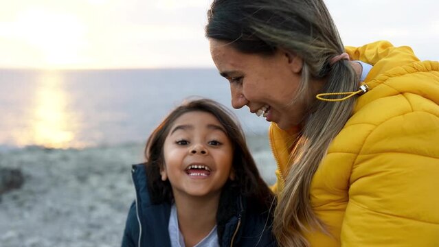 Happy latin mother and daughter smiling on camera with beach in the background during winter time	
