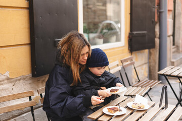 Happy mother hugs her son, a little good boy hold plate with cook. The family rest in the cafe, they eat desserts, laugh and play, happy family day. Selective focus. Woman look at boy, good mood.
