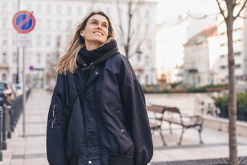 Happy blonde woman wear navy blue bomber jacket walking in the street and smiling, she look up....