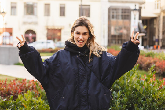 Cool blonde woman in navy blue bomber shows sign v, peace and friendship with one hand , open mouth and posing outside. Happy woman walk and posing outdoor in the city.