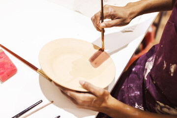  Closeup of a creative female hands skillfully paint a clay dish with paintbrush in an artisan workshop.