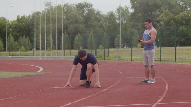 Young caucasian athlete starts abruptly from a low-start position while his asian trainer controls the time at the running track in an outdoor stadium on a cloudy day