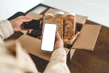 Phone with an isolated screen and credit card in hands against the background of a parcel with granola