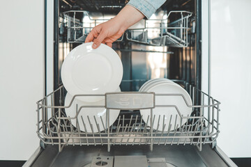 Female hand loading dished, empty out or unloading dishwasher with utensils. Kitchen appliances, lifestyle view. Woman puts a plate in the dishwasher or takes from it. Housewife does her housework