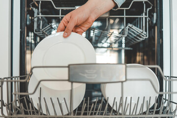 Female hand loading dished, empty out or unloading dishwasher with utensils. Kitchen appliances, lifestyle view. Woman puts a plate in the dishwasher or takes from it. Housewife does her housework