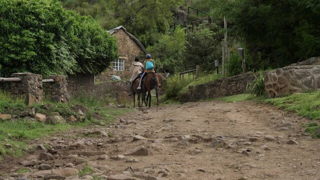 Africans trot horses on rocky path past stone house in Lesotho village