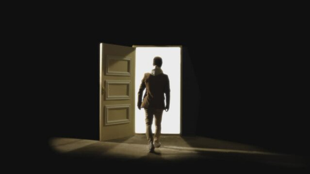 Back , rear view of man walking in front of an open door with darkness behind . Footage of person walking on floor . White light inside the door. Concept of anonymous,  spirit in coma or teleportation