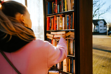 Young caucasian woman choosing books at free open public library at city park for book sharing...