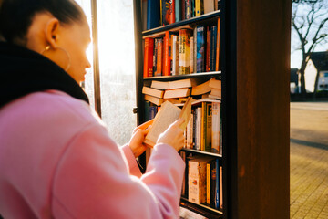 Young caucasian woman choosing books at free open public library at city park for book sharing...