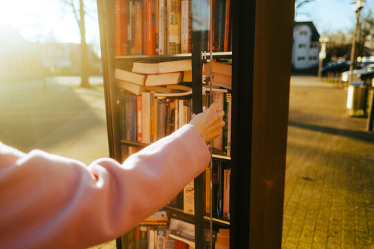 Young caucasian woman choosing books at free open public library at city park for book sharing among readers.
