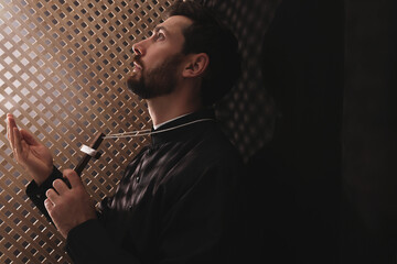 Catholic priest in cassock holding cross and praying to God in confessional - obrazy, fototapety, plakaty