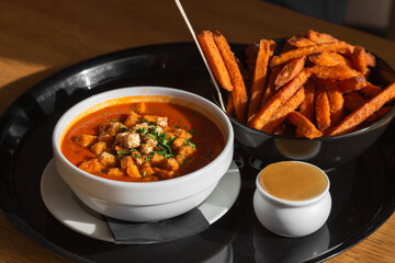 Bowl of red tomato soup gazpacho with croutons, vegetables and herbs, sweet potato fries with chili sauce on black tray.