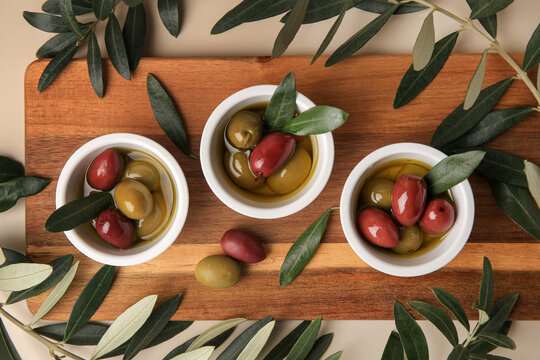Bowls with different ripe olives and leaves on beige background, flat lay