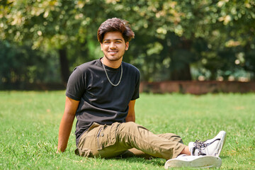 Smiling indian man candid portrait in black t shirt and silver neck chain sitting on green lawn in...