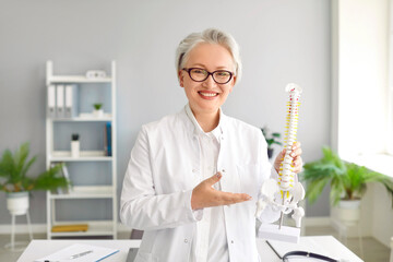 Happy doctor shows anatomical model of human spine at clinic. Portrait of cheerful middle aged...