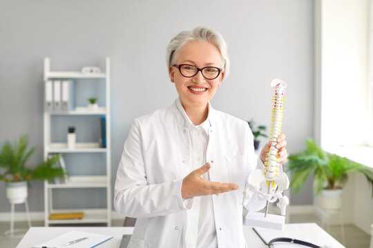 Happy Doctor Shows Anatomical Model Of Human Spine At Clinic. Portrait Of Cheerful Middle Aged Woman In White Medical Lab Coat And Glasses Holding Orthopedic Backbone Model And Smiling. Health Concept