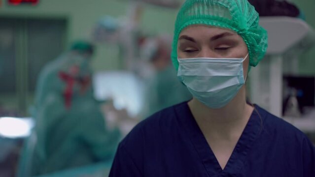 Portrait Of Serious Nurse Posing In Slow Motion In Operating Room And Looking Back At Colleagues Performing Surgery. Young Caucasian Woman Looking At Camera Standing In Hospital Indoors