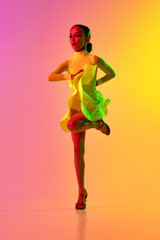 Elegant little girl in adorable stage outfit, dress dancing ballroom dance over gradient pink-yellow background in neon light filter. Concept of beauty, professional dances, skills