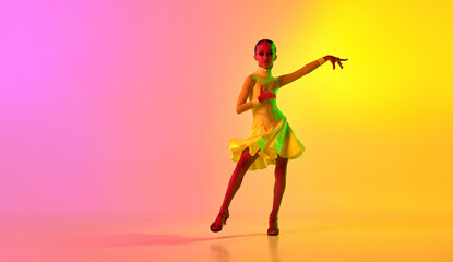 Elegant little girl in adorable stage outfit, dress dancing ballroom dance over gradient pink-yellow background in neon light filter. Concept of beauty, professional dances, skills