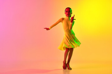 Emotional little girl in yellow stage dress dancing classical ballroom dance over gradient pink-yellow background in neon light filter.