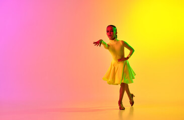 Fototapeta na wymiar Emotional little girl in yellow stage dress dancing classical ballroom dance over gradient pink-yellow background in neon light filter.