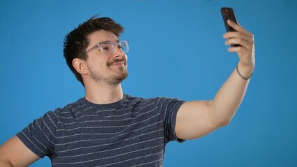 Fun young brunet man 20s wears striped t-shirt purple shirt doing selfie shot on mobile cell phone post photo on social network meet greet wave hand isolated on blue background studio portrait