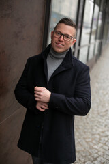 Handsome smiling man in glasses and black coat near building. Portrait of business man walking outdoors in cold day.