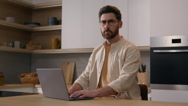 40s serious Caucasian man renter homeowner male browsing laptop at kitchen buying food online shopping looking at camera. Freelancer businessman working from home with computer service posing at table