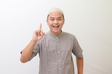 Portrait of a young Asian man wearing a Muslim shirt and hat smiling broadly with joy and raising his index finger upwards. Combed image on white background 