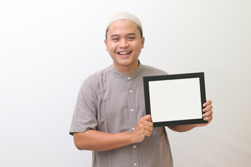 The portrait of the religious wearing a muslim gray shirt and a white hat smiled broadly holding a whiteboard. Isolated image on white background