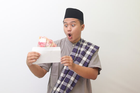 
Portrait of asian man wearing black hat and gray muslim shirt happy holding envelope filled with money with surprised expression looking at money, isolated image on gray background 