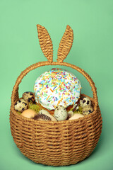 Easter decorations in a basket with rabbit ears. Easter cake and eggs on moss, quail eggs and feathers on mint green background.