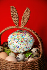Easter celebration. Easter decorations in a basket with rabbit ears. Easter cake and eggs on moss, quail eggs and feathers on a red background. Traditions and religion.