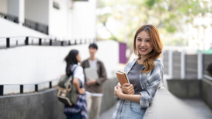 Cute asian girl student with hold a book and laptop near the campus against the background of a group of students.