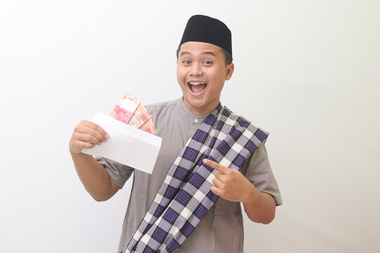 Portrait of smiling Asian man smiling happily at camera wearing gray muslim shirt and black hat with sarong slung over shoulder pointing to envelope filled with money he is carrying. Isolated image on