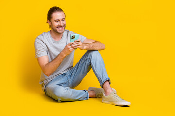 Full length photo of good mood optimistic guy dressed light t-shirt jeans look at smartphone chatting isolated on yellow color background