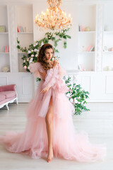 Young brunette girl with long beautiful hair in a lush pink tulle dress in a light white studio on the background of shelves with books, a fireplace and flower arrangements