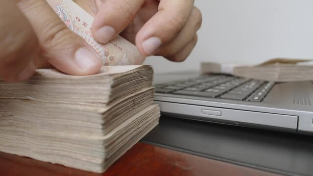 Select focus of hands counting a lot of thousands Thai baht banknotes money near laptop or notebook computer. The baht is the official currency of Thailand.