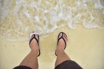 Close-up of a man in black sandals standing at the beach. with soft foamy waves underneath, top view, Thailand travel concept, Phuket