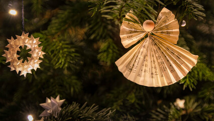 Ein Papierengel und ein Weihnachtsstern hängen an einem dezent beleuchteten Weihnachtsbaum