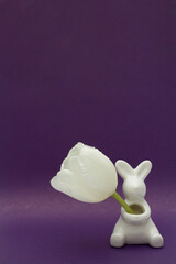 Vertical banner with space for text. Ceramic figurine of an cute Easter bunny with white tulip flower on dark purple background. Happy Easter religious holiday greeting card. Copyspace. Celebration