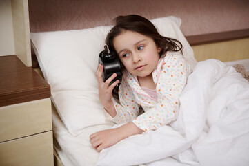 Charming European little girl holding an alarm clock near her face, dressed in glamorous pajamas, getting ready for bed. Daily routines in the childhood. The concept of relaxing, bedtime and waking up