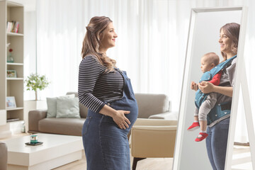 Pregnant woman looking herself at a mirror holding a baby at home